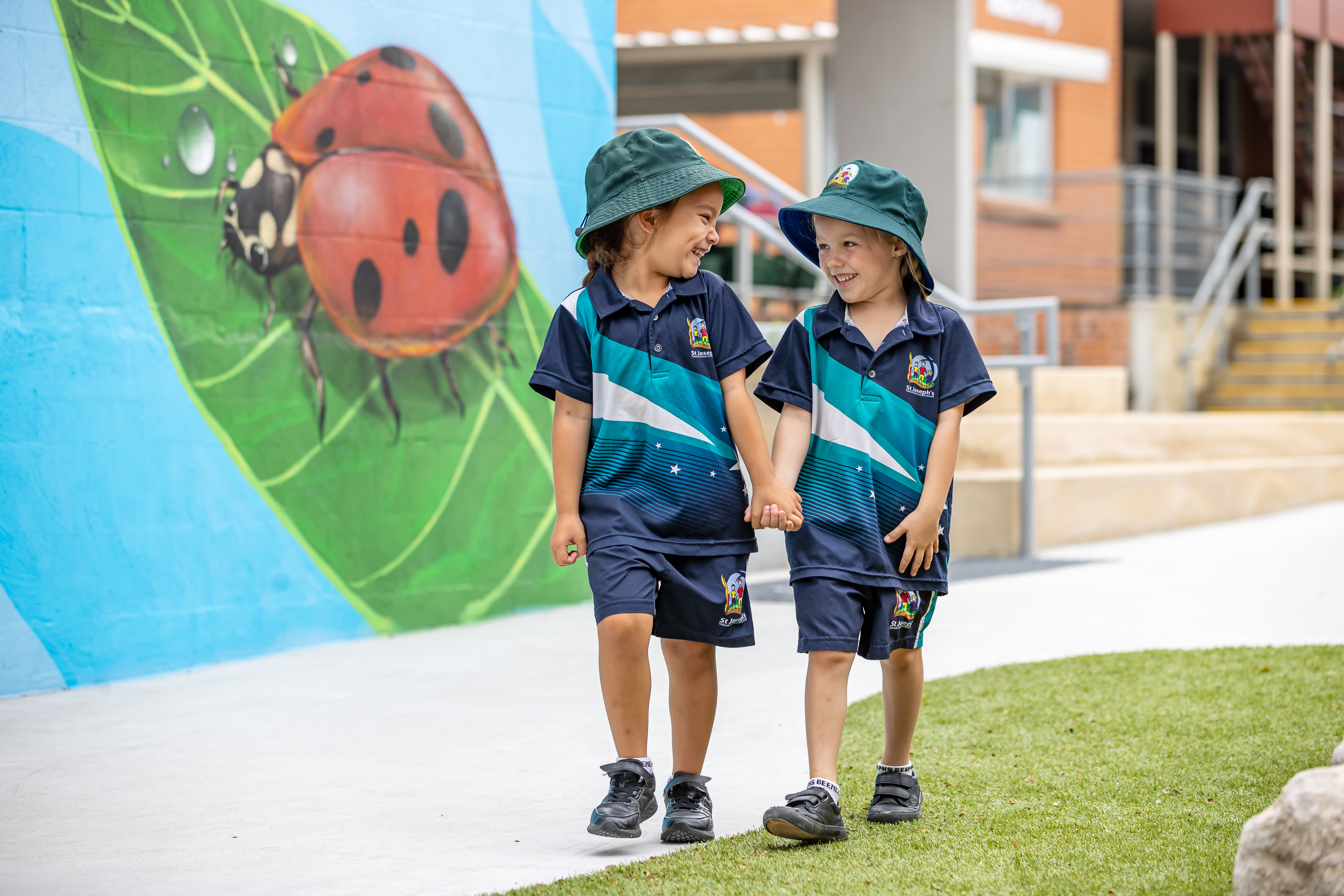 Two children in school uniforms walking hand in hand past a large mural of a ladybird on a wall outdoors.