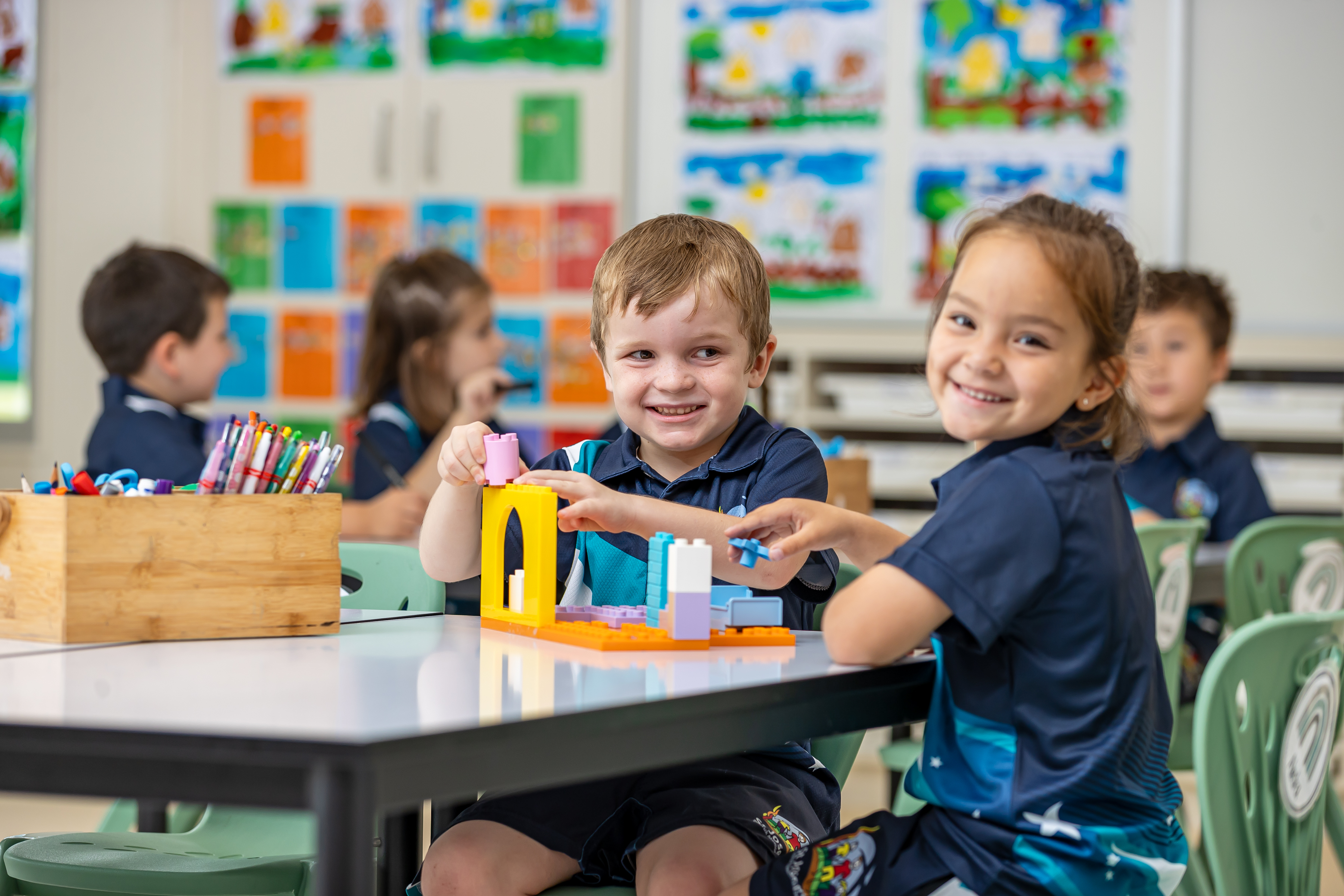 Children at a classroom table building with colourful blocks, with drawings displayed on the wall behind them.