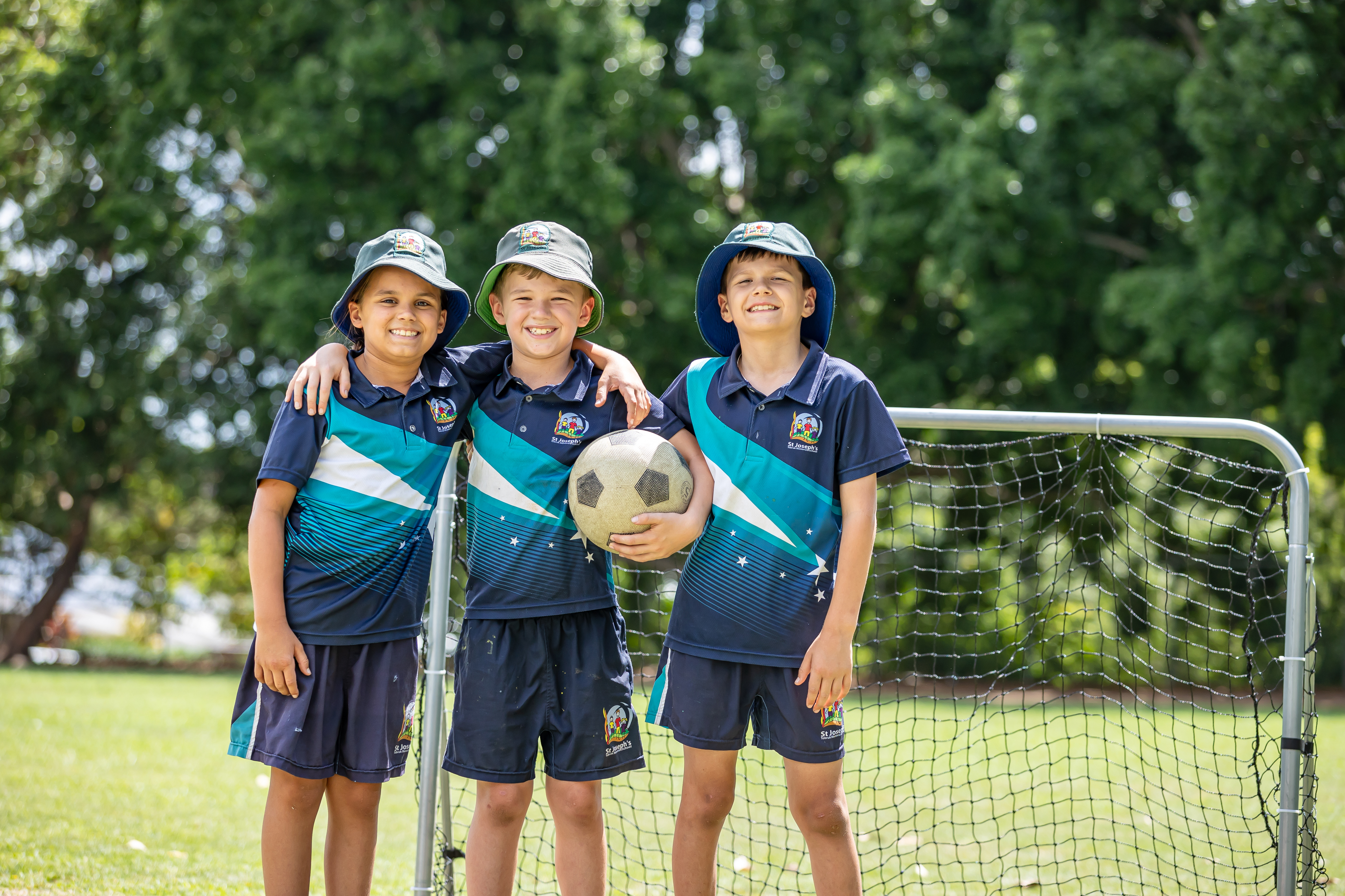 Three students in sports uniforms standing in front of a goal, with one holding a soccer ball outdoors.