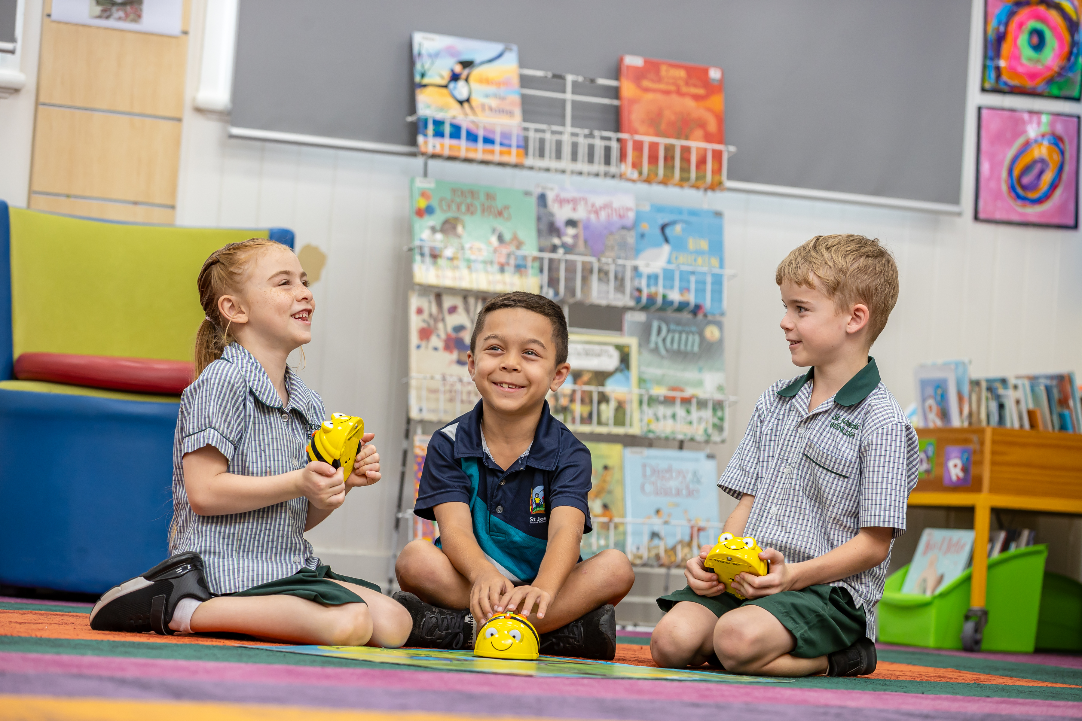 Three children sitting on a classroom rug holding small yellow devices during a learning activity, with books displayed on shelves behind them.