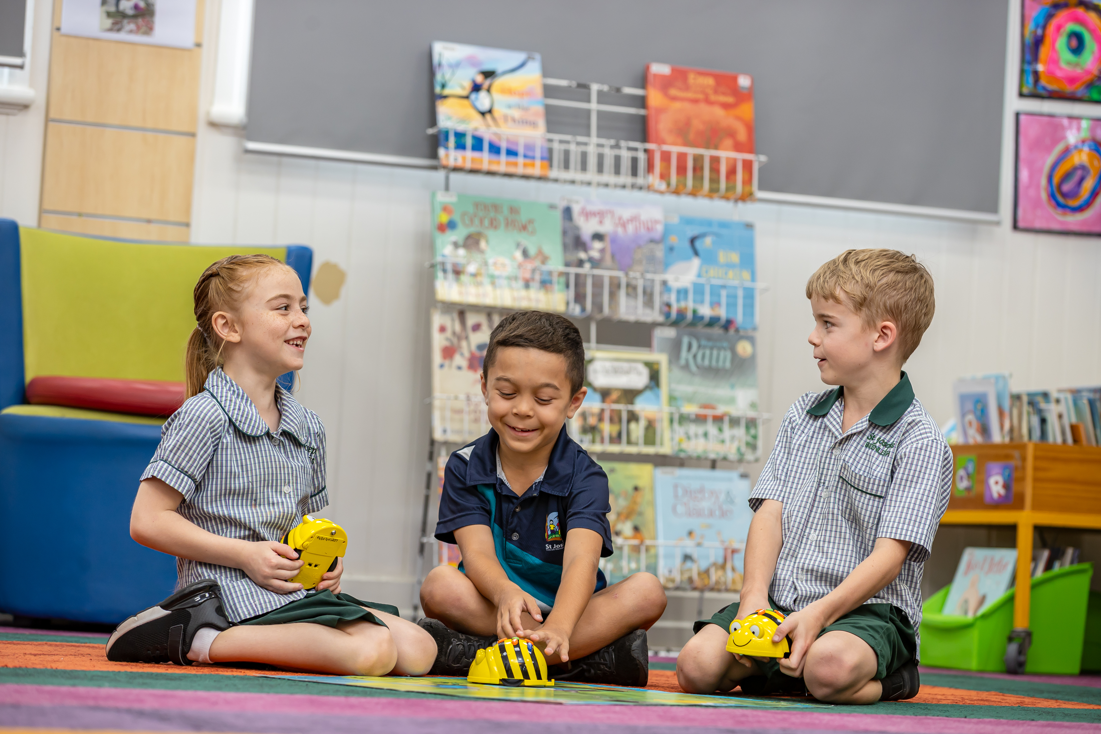 Three children sitting on a classroom rug using small yellow robotic devices during a learning activity, with bookshelves in the background.