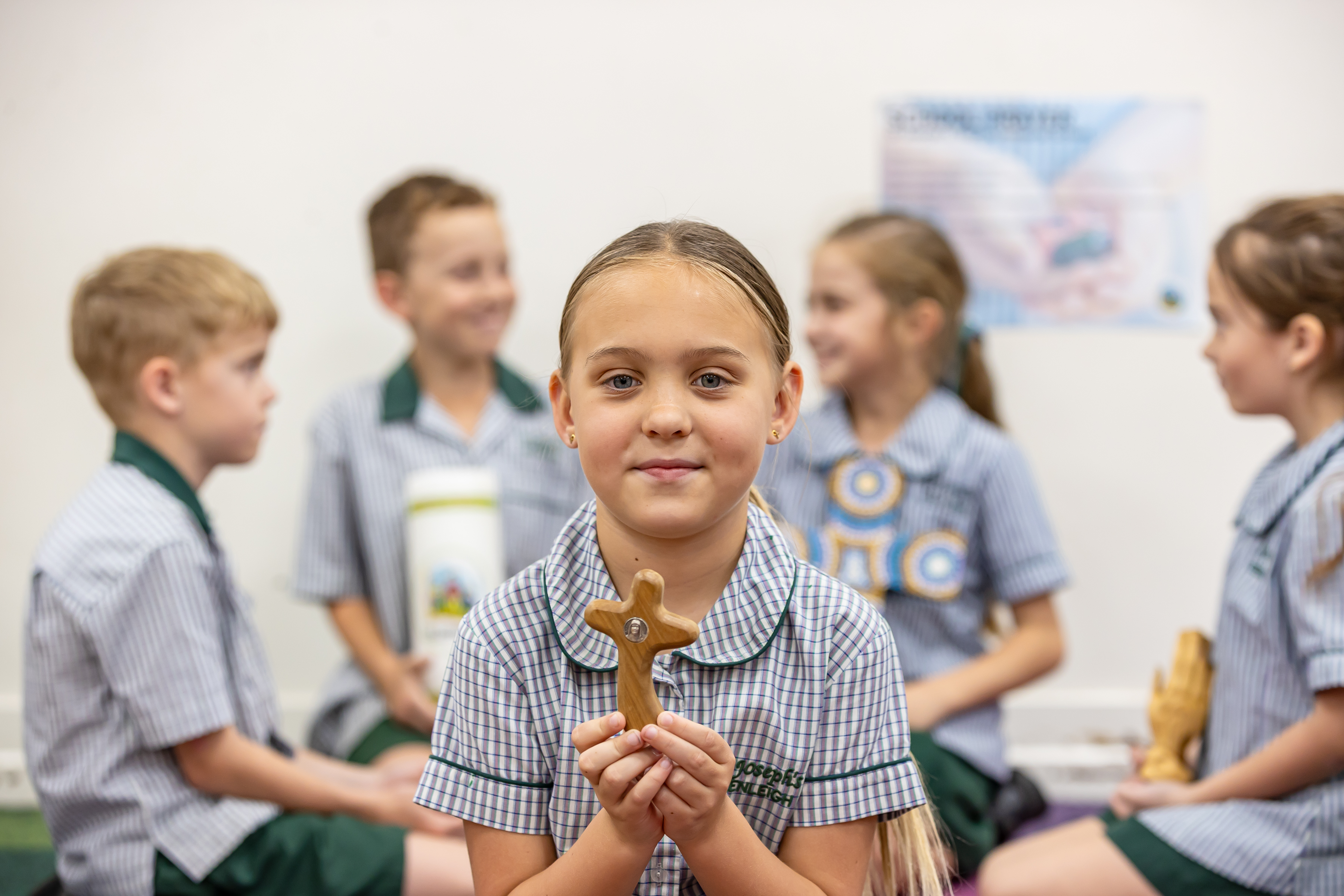 Child holding a wooden cross in a classroom, with other students in uniform in the background.